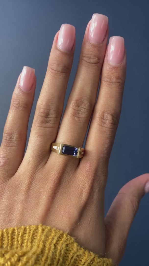 andmade yellow gold ring featuring a vibrant emerald-cut tanzanite gem with baguette diamonds on the sides and a matte satin finish